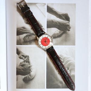 May include: A wristwatch with a silver case and a red and white face. The watch has a black leather band with red stitching. The watch is displayed on a page with black and white photographs.