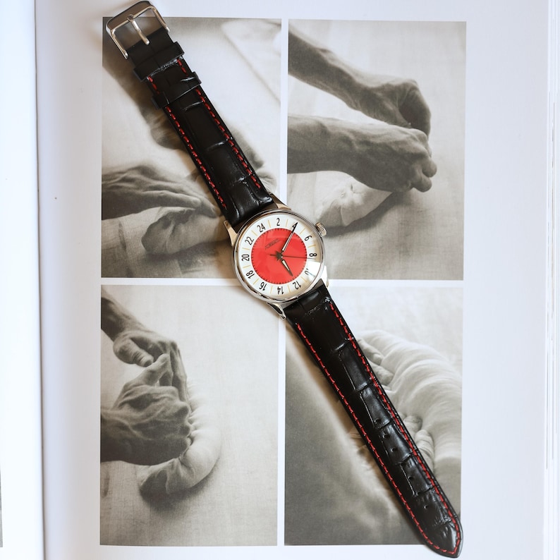 May include: A wristwatch with a silver case and a red and white face. The watch has a black leather band with red stitching. The watch is displayed on a page with black and white photographs.