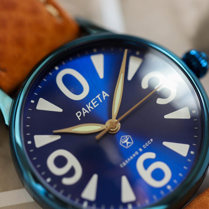 May include: Close-up of a blue wristwatch with a dark blue face. The watch face features white numerals and the word "PAKETA" in white. The watch has gold-coloured hands and a brown leather strap.