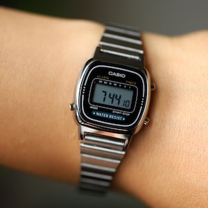 K&ouml;nnte beinhalten: Eine silberfarbene Digitalarmbanduhr mit einem rechteckigen schwarzen Zifferblatt, das das Casio-Logo, Alarm- und Timer-Funktionen sowie den Schriftzug Water Resist zeigt. Die Uhr hat ein Metallgliederarmband und eine Digitalanzeige mit 7:44:10 Uhr.
