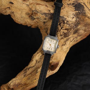 May include: A square-faced wristwatch with a black leather band. The watch face has white Roman numeral hour markers and black hands.