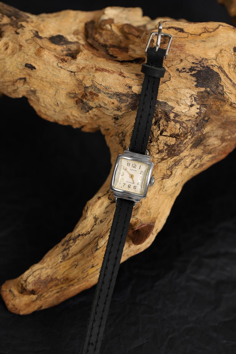 May include: A square-faced wristwatch with a black leather band. The watch face has white Roman numeral hour markers and black hands.