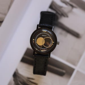 May include: A black wristwatch with a round face featuring gold accents and the word "PAKETA". The watch has a black leather band. The watch face has a gold circle and a gold ring design.