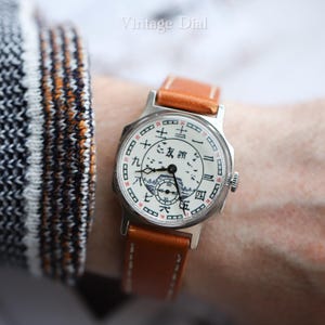 Puede incluir: Un reloj de pulsera vintage con una esfera blanca con caracteres chinos negros y rojos y una pequeña ilustración de paisaje. El reloj tiene una caja plateada y una correa de cuero marrón. El texto "Vintage Dial" está en la parte superior.