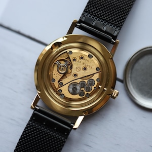 May include: Close-up of a gold-toned watch with the back removed, revealing the intricate mechanical movement. The watch has a black mesh band. The internal gears and components are visible, showcasing the watch's craftsmanship.