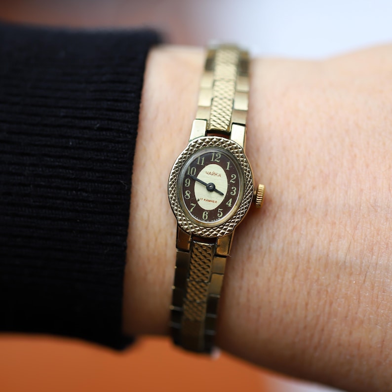 Puede incluir: Un reloj de pulsera vintage dorado con una correa texturizada y una esfera ovalada. La esfera del reloj es marr&oacute;n con n&uacute;meros y agujas doradas. La palabra "Чайка" est&aacute; impresa en la esfera.