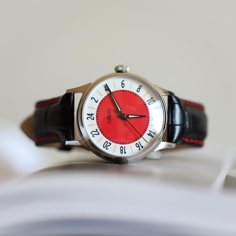 May include: A vintage wristwatch with a silver case, a red dial, and a black leather band. The watch face features white numerals and hands, with a 24-hour time display. The band has red stitching.