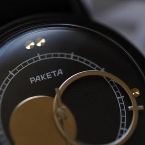 May include: Close-up of a black watch face with gold accents. The word "PAKETA" is printed in white. The watch features a gold-colored circular element and a gold-colored hand. The watch face has a circular design with a series of small lines.