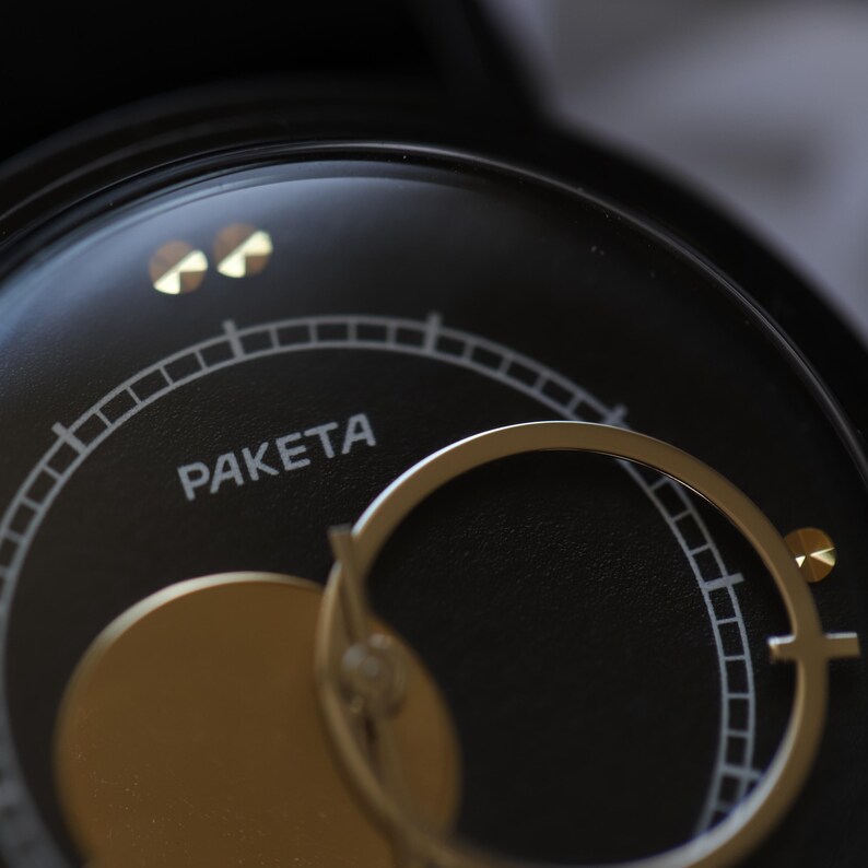May include: Close-up of a black watch face with gold accents. The word "PAKETA" is printed in white. The watch features a gold-colored circular element and a gold-colored hand. The watch face has a circular design with a series of small lines.