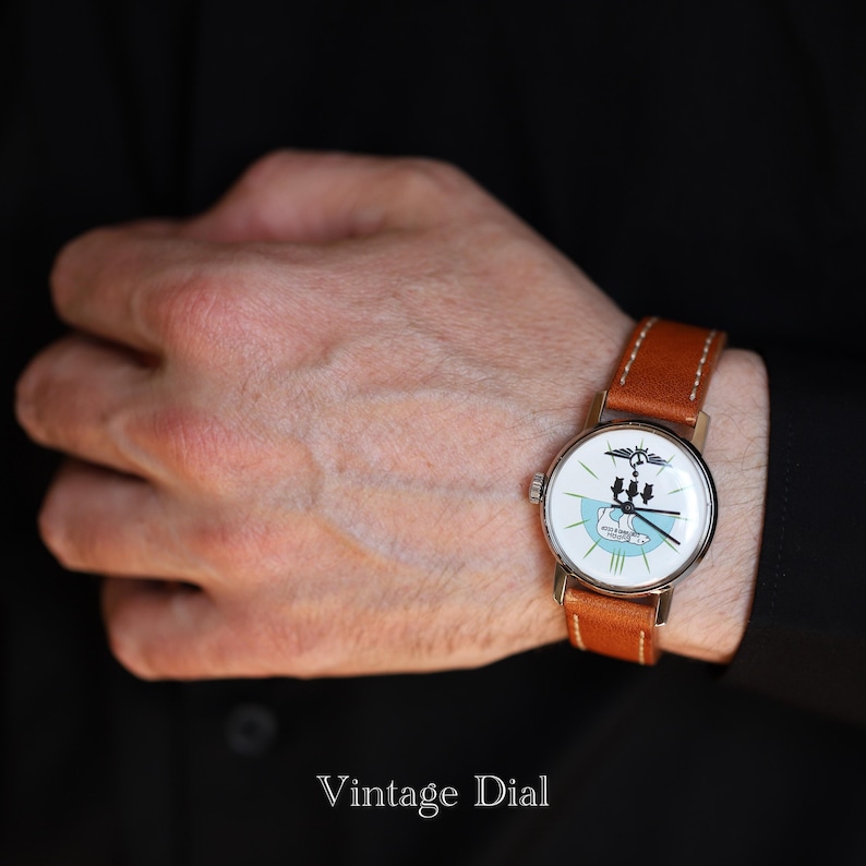 Puede incluir: Un reloj de pulsera vintage con esfera blanca, un emblema negro y azul, y agujas negras. Tiene una correa de cuero marr&oacute;n. El texto "Vintage Dial" est&aacute; en la parte inferior.