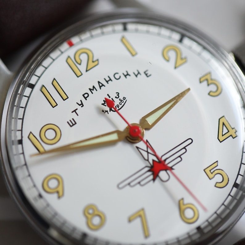 May include: Close-up of a vintage wristwatch with a white face and gold numerals. The watch features gold hands, a red second hand, and a black and red emblem. The watch face has the text "Штурманские".