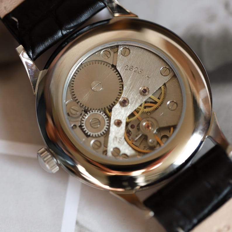 May include: Close-up of a vintage wristwatch's back, showcasing the intricate mechanical movement. The watch has a silver-toned case and a black leather strap. The internal gears and components are visible through the clear back, with the number "2623.4" etched on a metal plate.