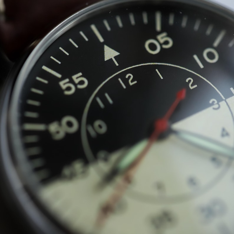 May include: Close-up of a black stopwatch with white numerals and a red second hand. The watch face displays the numbers 05, 10, 12, 50, and 55. The watch has a white arrow and a white and silver dial.