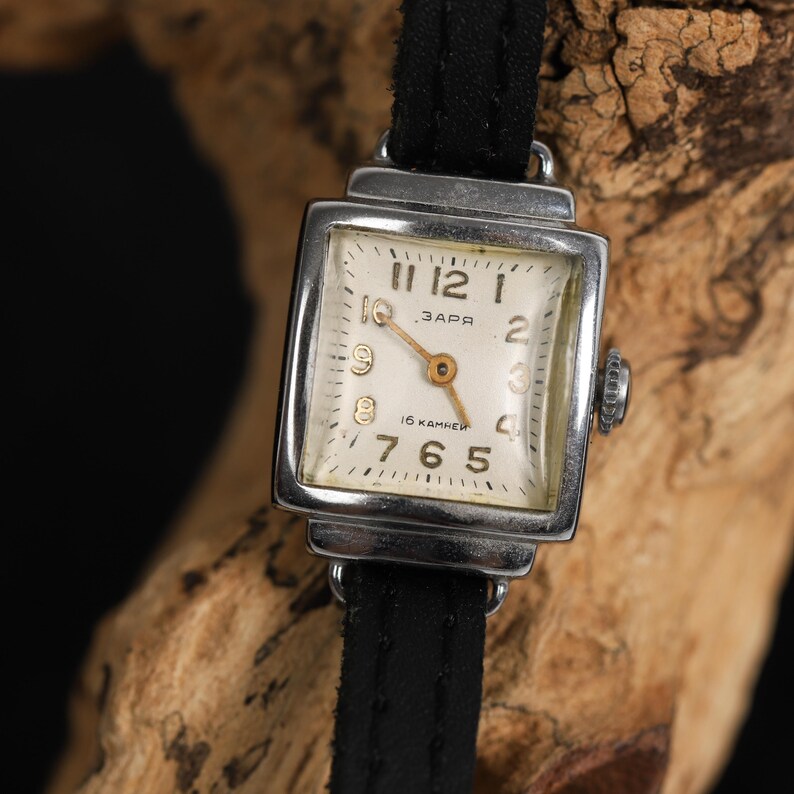 May include: A silver square wristwatch with a white face and black leather band. The watch face has black Roman numeral hour markers, black minute markers, and a gold second hand. The watch face also has the text "ЗАРЯ" and "16 КАМЕНЕЙ".