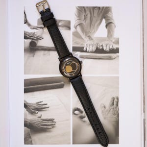 May include: Black wristwatch with a textured leather band and a gold-toned face. The watch has a round black dial with gold accents. The watch is displayed on a black and white photo collage.