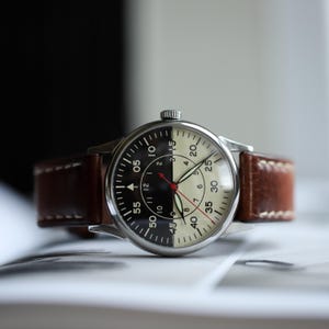 May include: A wristwatch with a two-tone black and cream face, featuring a brown leather strap. The watch has a silver-colored case and a red second hand. The face displays numbers and markers for timekeeping.