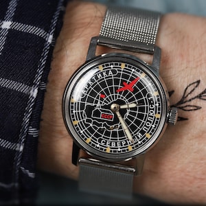 May include: A black and white wristwatch with a silver metal band. The watch face features a compass design with a red airplane and the text "АРКТИКА СЕВЕРНЫЙ ПОЛЮС-1" in Cyrillic script. The watch face also includes the text "CCCP" in Cyrillic script.