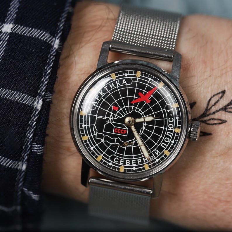 May include: A black and white wristwatch with a silver metal band. The watch face features a compass design with a red airplane and the text "АРКТИКА СЕВЕРНЫЙ ПОЛЮС-1" in Cyrillic script. The watch face also includes the text "CCCP" in Cyrillic script.