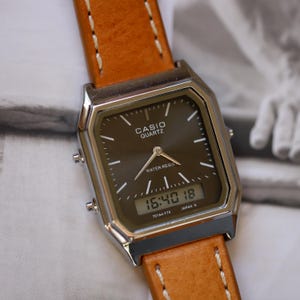 May include: A Casio Quartz watch with a brown face and a digital display showing the time. The watch has a silver-colored case and a light brown leather strap with white stitching. The watch is water resistant.