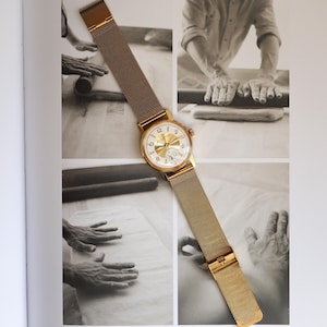 K&ouml;nnte beinhalten: Eine goldfarbene Armbanduhr mit rundem Zifferblatt, wei&szlig;em Hintergrund und goldfarbenen arabischen Ziffern. Die Uhr verf&uuml;gt &uuml;ber ein goldfarbenes Metall-Mesh-Armband mit Schiebeverschluss. Das Zifferblatt hat ein Hilfszifferblatt bei sechs Uhr.