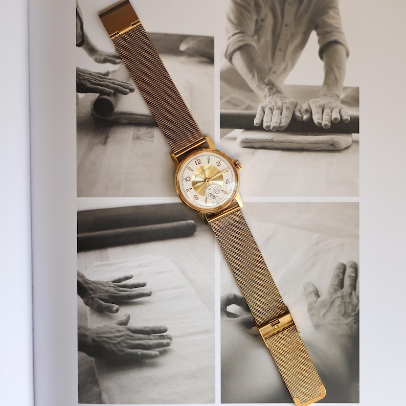 K&ouml;nnte beinhalten: Eine goldfarbene Armbanduhr mit rundem Zifferblatt, wei&szlig;em Hintergrund und goldfarbenen arabischen Ziffern. Die Uhr verf&uuml;gt &uuml;ber ein goldfarbenes Metall-Mesh-Armband mit Schiebeverschluss. Das Zifferblatt hat ein Hilfszifferblatt bei sechs Uhr.