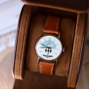 Puede incluir: Un reloj de pulsera vintage con una esfera blanca con un gr&aacute;fico de ping&uuml;inos y texto. El reloj tiene una correa de cuero marr&oacute;n y una caja plateada. El reloj se muestra en un estuche de cuero marr&oacute;n.