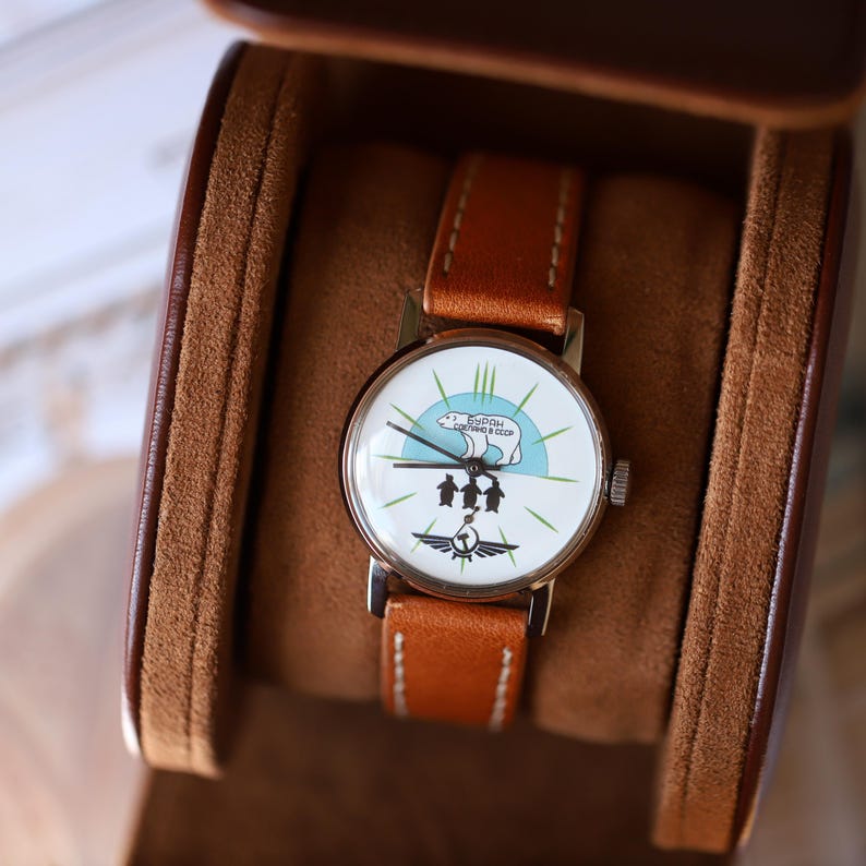 Puede incluir: Un reloj de pulsera vintage con una esfera blanca con un gr&aacute;fico de ping&uuml;inos y texto. El reloj tiene una correa de cuero marr&oacute;n y una caja plateada. El reloj se muestra en un estuche de cuero marr&oacute;n.