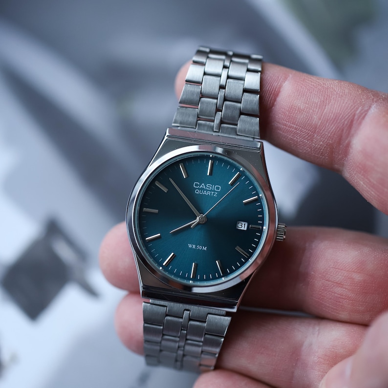 May include: A silver-toned Casio Quartz watch with a teal face and a date display at the 3 o'clock position. The watch has a stainless steel band and is held by a hand.