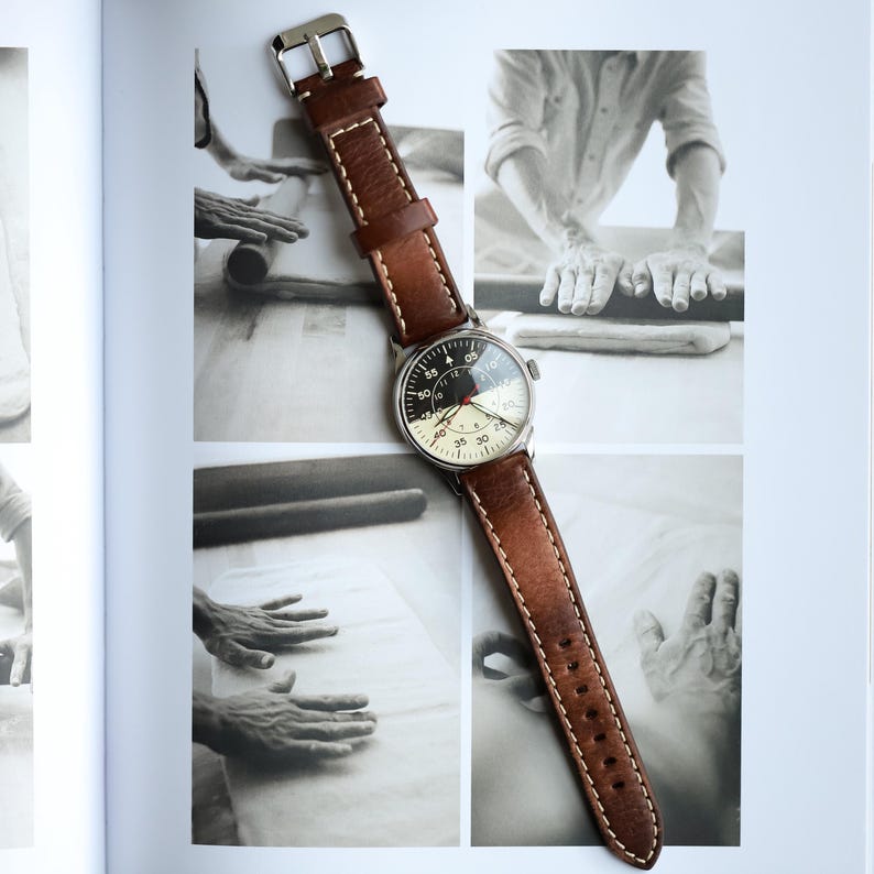 May include: A wristwatch with a brown leather band and a silver-colored case. The watch face is off-white with black and red details. The band is stitched with white thread. The watch is displayed on a book with black and white photos.