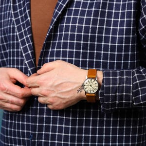 May include: A man wearing a blue and white plaid shirt with a brown leather wristwatch. The watch has a white face with black markings and a silver case.