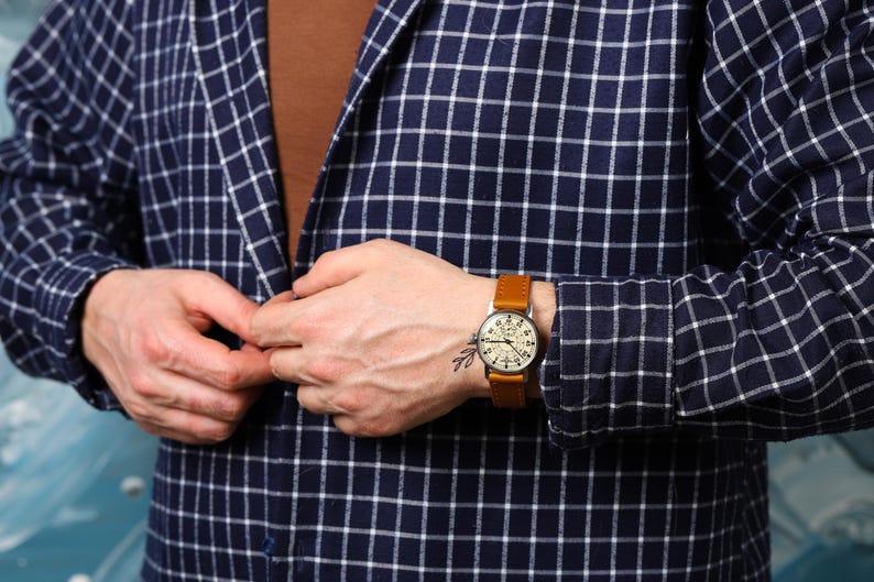 May include: A man wearing a blue and white plaid shirt with a brown leather wristwatch. The watch has a white face with black markings and a silver case.