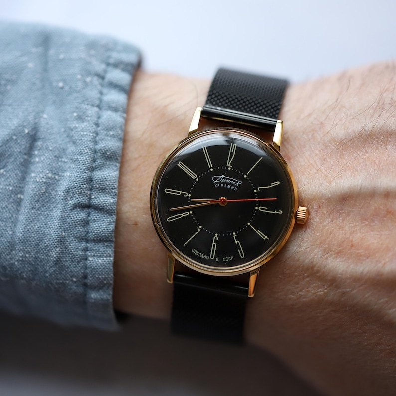 May include: A vintage gold-toned wristwatch with a black face and a black mesh band. The watch face features gold-colored hour markers and hands, with the text "23 KAMHЯ" and "СДЕЛАНО В СССР".