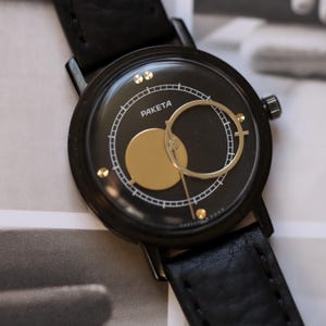 May include: Black wristwatch with a round face featuring the word "PAKETA" in white. The watch face has gold-colored accents and a black leather band. The watch has a minimalist design.
