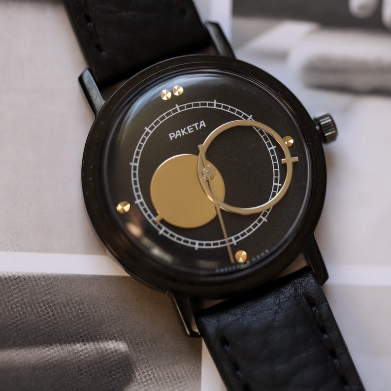 May include: Black wristwatch with a round face featuring the word "PAKETA" in white. The watch face has gold-colored accents and a black leather band. The watch has a minimalist design.