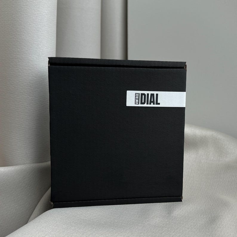 May include: A black square box with the word "DIAL" printed on a white label. The word "VINTAGE" is printed vertically on the left side of the label. The box is sitting on a light-colored surface.