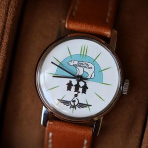 Puede incluir: Un reloj de pulsera vintage con una esfera blanca que presenta un oso polar, ping&uuml;inos y el texto "БУРАН СДЕЛАНО В СССР". El reloj tiene una caja plateada y una correa de cuero marr&oacute;n. La esfera tiene agujas negras y detalles en verde.