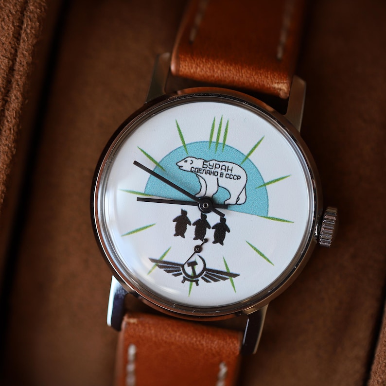 Puede incluir: Un reloj de pulsera vintage con una esfera blanca que presenta un oso polar, ping&uuml;inos y el texto "БУРАН СДЕЛАНО В СССР". El reloj tiene una caja plateada y una correa de cuero marr&oacute;n. La esfera tiene agujas negras y detalles en verde.