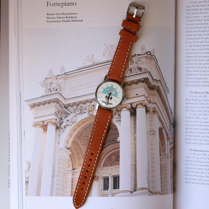 Puede incluir: Un reloj de pulsera con una correa de cuero marr&oacute;n claro y una caja plateada. La esfera del reloj presenta un dise&ntilde;o azul y blanco. El reloj est&aacute; sobre un libro abierto con im&aacute;genes arquitect&oacute;nicas.