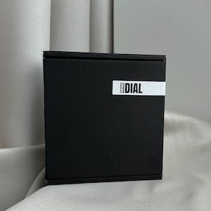 May include: A black square box with a white label that reads "VINTAGE DIAL". The box is made of a matte material and has a simple, clean design. The background is a neutral gray.
