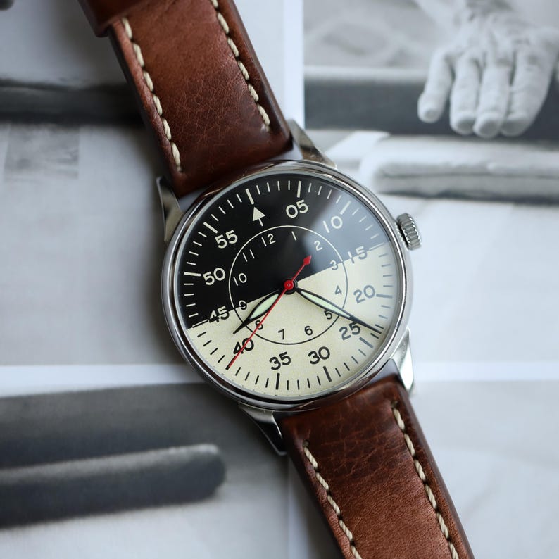 May include: A vintage-style wristwatch with a black and cream-colored face and a brown leather strap. The watch face features black numerals and a red second hand. The strap is brown leather with white stitching.