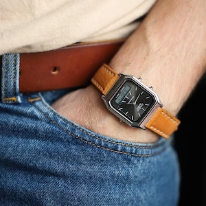 May include: A silver-toned digital watch with a black face and a tan leather band. The watch is on a wrist, with a brown leather belt and blue jeans in the background. The watch displays the time.