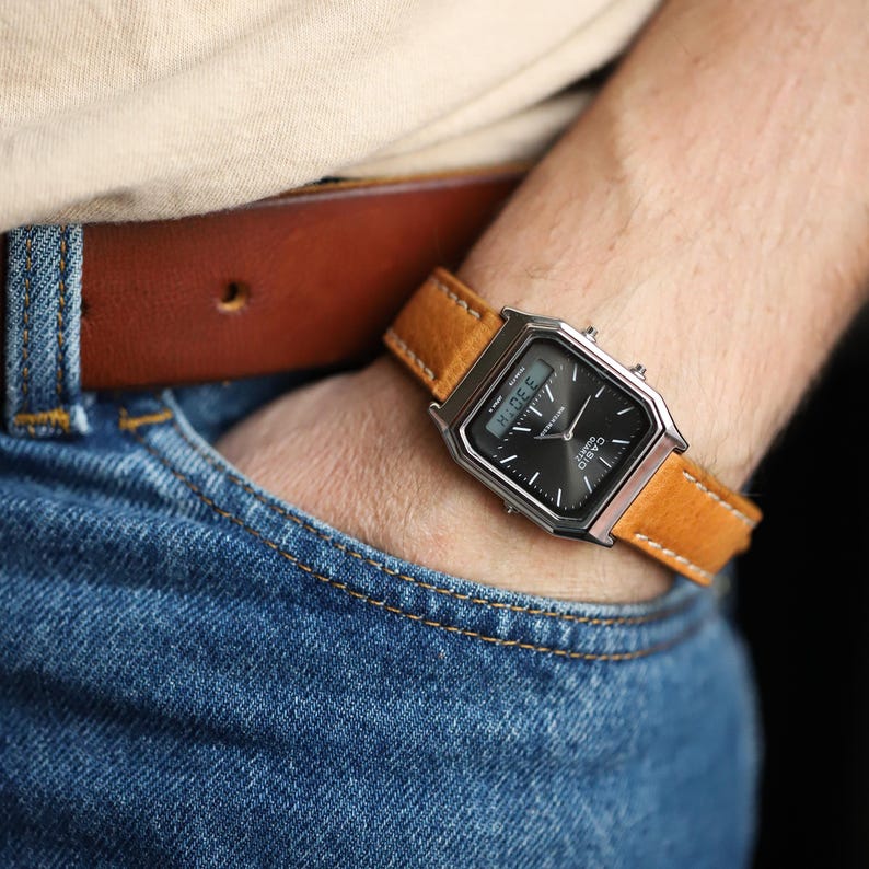 May include: A silver-toned digital watch with a black face and a tan leather band. The watch is on a wrist, with a brown leather belt and blue jeans in the background. The watch displays the time.