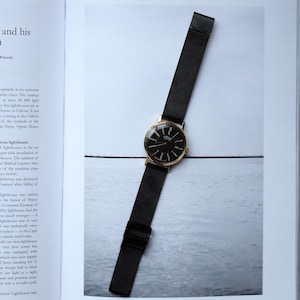 May include: A gold-toned wristwatch with a black face and hands, and a black mesh band. The watch is displayed on a light-colored surface, showcasing its elegant design. The watch face has the word "ORION" on it.