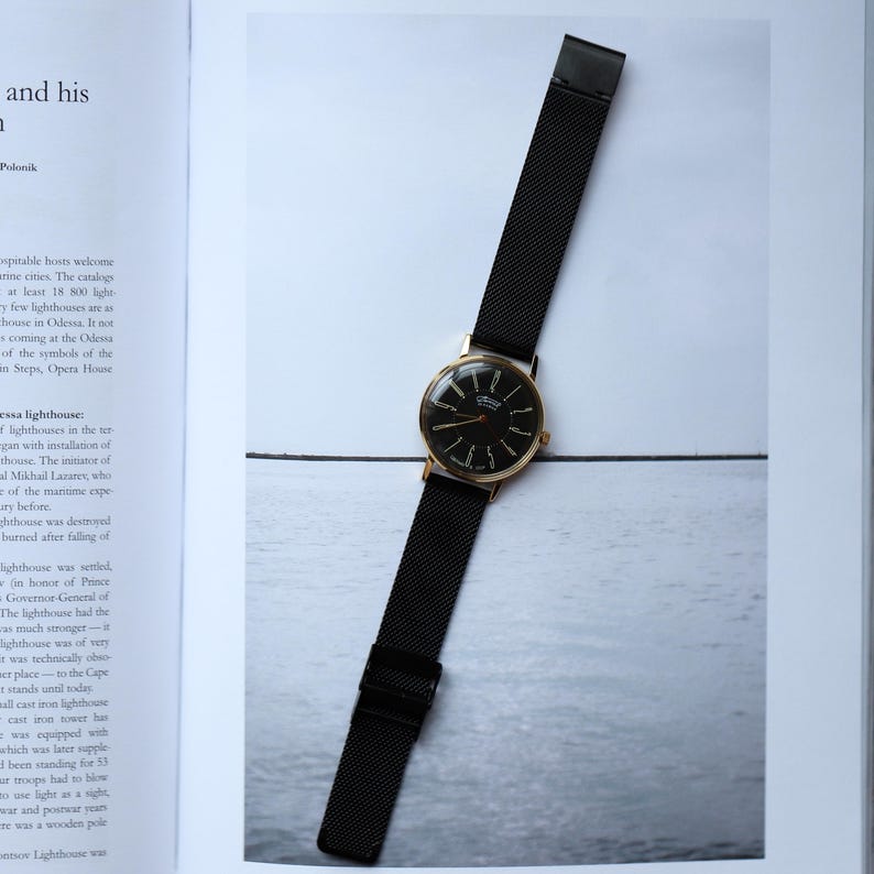 May include: A gold-toned wristwatch with a black face and hands, and a black mesh band. The watch is displayed on a light-colored surface, showcasing its elegant design. The watch face has the word "ORION" on it.