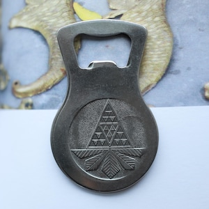 Könnte beinhalten: Ein silberfarbener Flaschenöffner mit geometrischem Design. Der Öffner hat ein kreisförmiges Emblem mit einem Dreiecksmuster und einem stilisierten Blattdesign am unteren Rand. Er hat einen gebogenen Griff und einen Flaschenverschlussentferner.