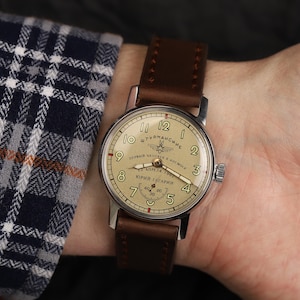 Pode incluir: Um rel&oacute;gio de pulso prateado com uma pulseira de couro marrom. O mostrador do rel&oacute;gio &eacute; de cor creme com marcadores de hora romanos pretos e marcadores de minutos pretos. O mostrador do rel&oacute;gio tamb&eacute;m apresenta um pequeno gr&aacute;fico de avi&atilde;o e o texto "ШТУРМАНСКИЕ ПЕРВЫЙ ЧЕЛОВЕК В КОСМОС 12 АПРЕЛЯ 1961 ЮРИЙ ГАГАРИН".