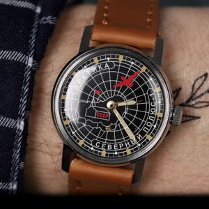 May include: A black and white wristwatch with a compass face. The watch face features a red airplane, the text "АРКТИКА СЕВЕРНЫЙ ПОЛЮС-1", and the abbreviation "CCCP". The watch has a brown leather band.