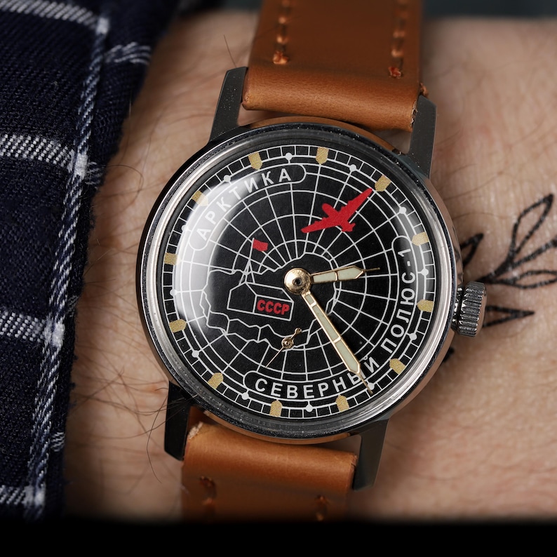 May include: A black and white wristwatch with a compass face. The watch face features a red airplane, the text "АРКТИКА СЕВЕРНЫЙ ПОЛЮС-1", and the abbreviation "CCCP". The watch has a brown leather band.