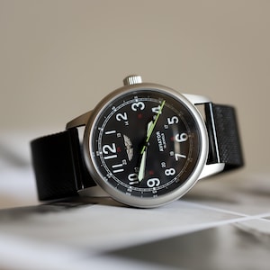 Puede incluir: Un reloj de pulsera redondo, plateado, con esfera negra y correa de tela negra. La esfera muestra números blancos, un pequeño logotipo y el texto "AVIATOR 19 JEWELS". Las manecillas son blancas y verde neón.