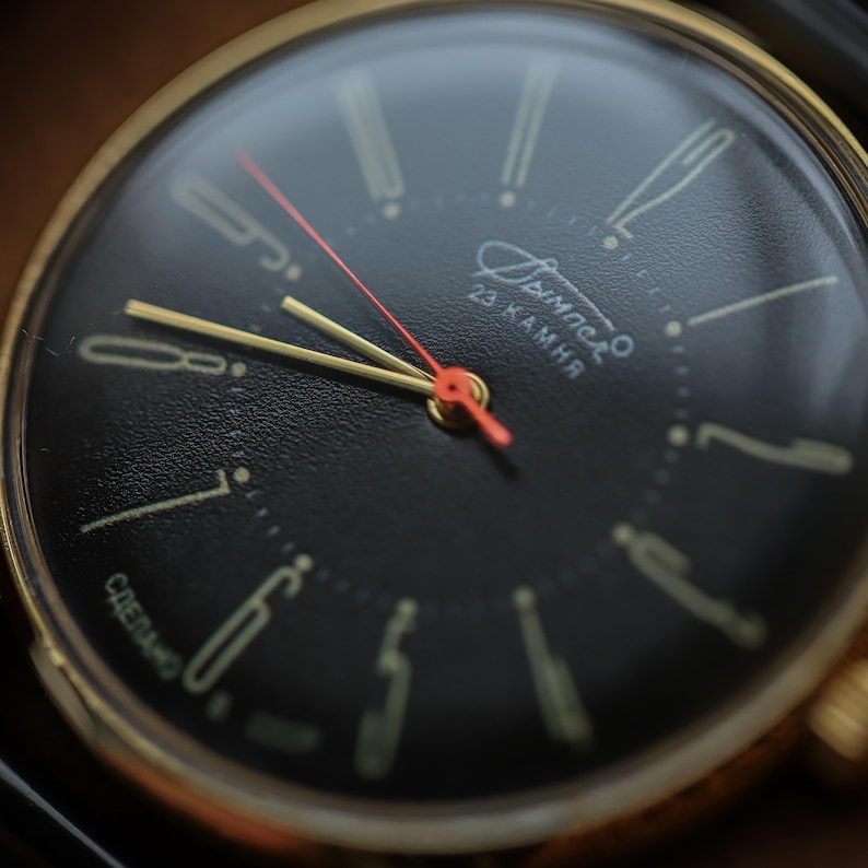 May include: Close-up of a vintage wristwatch with a black face and gold-colored hands and casing. The watch features a red second hand and gold-colored hour markers. The watch face has the text "Димacкo" and "23 камня".